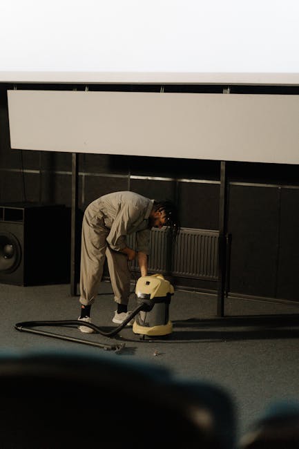 A person dressed in light-colored work overalls is kneeling on a carpeted floor, operating a yellow industrial vacuum cleaner used for deep cleaning or surface sanitisation. The room features dark wall panels and a white horizontal panel above a heating vent, with additional equipment such as a speaker in the background. Bright, even lighting illuminates the space, emphasizing its cleanliness and the professional cleaning process. This scene reflects a comprehensive approach to commercial or domestic surface cleaning, consistent with services offered by Finchley Cleaner as promoted on the East Finchley Carpet Cleaning Same Day Service page.