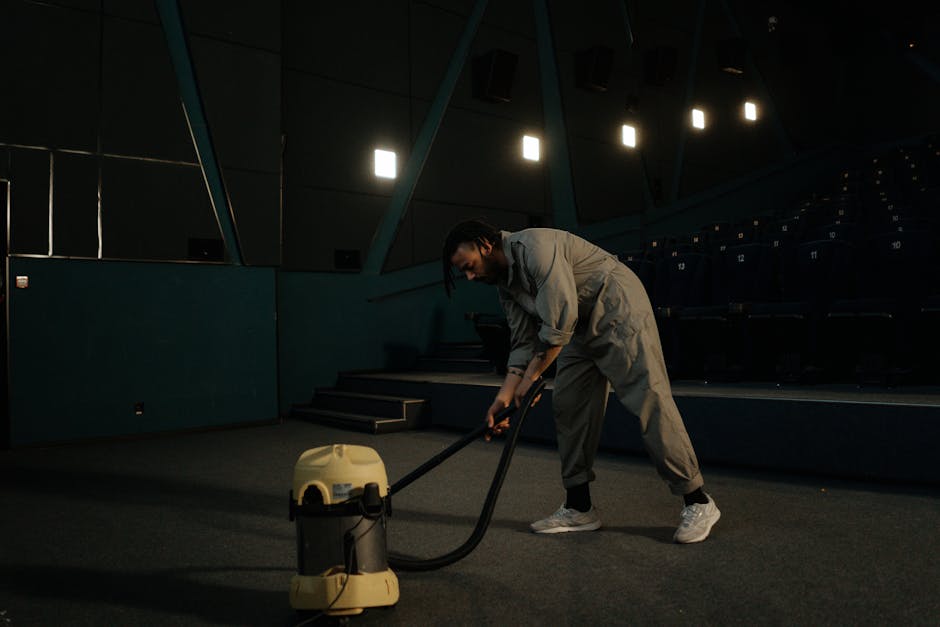A person dressed in light-colored work overalls is kneeling on a carpeted floor, operating a yellow industrial vacuum cleaner used for deep cleaning or surface sanitisation. The room features dark wall panels and a white horizontal panel above a heating vent, with additional equipment such as a speaker in the background. Bright, even lighting illuminates the space, emphasizing its cleanliness and the professional cleaning process. This scene reflects a comprehensive approach to commercial or domestic surface cleaning, consistent with services offered by Finchley Cleaner as promoted on the East Finchley Carpet Cleaning Same Day Service page.