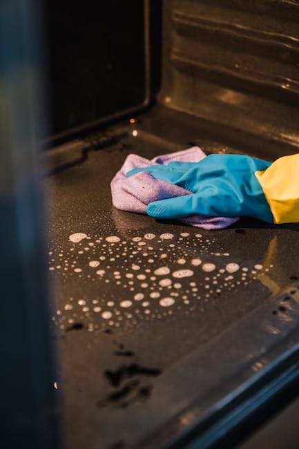 Close-up view of a black oven interior surface being cleaned by a person wearing bright blue cleaning gloves and holding a purple microfiber cloth. The oven's ceramic or enamel surface shows soapy water droplets and foam, indicating an ongoing deep cleaning process. The surrounding oven walls are made of stainless steel, reflecting light from the interior. The countertop appears clean and free of debris, with the lighting emphasizing the shine and cleanliness of the oven interior. The scene highlights professional domestic or commercial oven sanitisation efforts, as promoted by Finchley Cleaner, with a focus on thorough surface cleaning to ensure hygiene and shine in kitchen appliances.