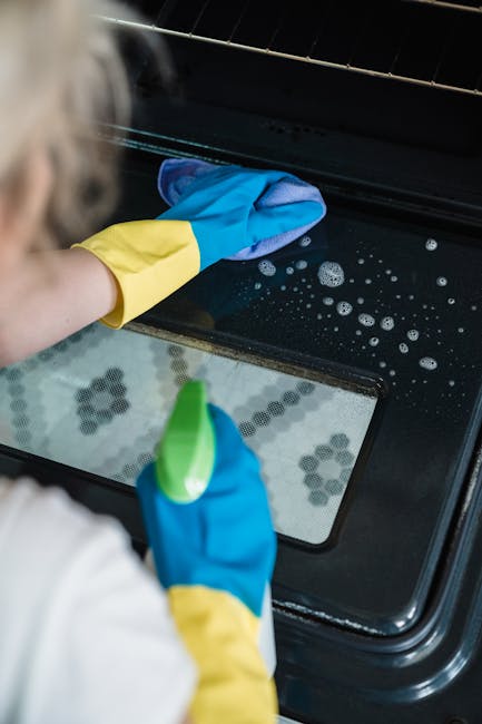 A person wearing blue and yellow rubber cleaning gloves is engaged in surface cleaning of the inside of an oven, focusing on the oven door and glass window. The oven appears to be part of a modern kitchen with a black ceramic or stainless steel appliance. The person is using a cloth or sponge to wipe the oven interior, and soap bubbles or cleaning foam are visible on the surface. The scene is well-lit, emphasizing the clean and shiny condition of the oven's surfaces. This image illustrates deep cleaning and sanitisation practices relevant to domestic cleaning, reflecting the expertise of Finchley Cleaner in maintaining hygienic kitchen appliances in residential settings.