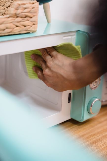 Close-up view of a black oven interior surface being cleaned by a person wearing bright blue cleaning gloves and holding a purple microfiber cloth. The oven's ceramic or enamel surface shows soapy water droplets and foam, indicating an ongoing deep cleaning process. The surrounding oven walls are made of stainless steel, reflecting light from the interior. The countertop appears clean and free of debris, with the lighting emphasizing the shine and cleanliness of the oven interior. The scene highlights professional domestic or commercial oven sanitisation efforts, as promoted by Finchley Cleaner, with a focus on thorough surface cleaning to ensure hygiene and shine in kitchen appliances.