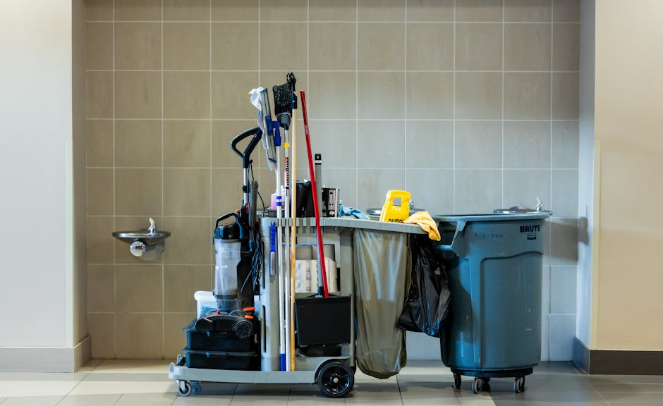 A cleaning trolley positioned against a beige tiled wall in a commercial space, holding various cleaning tools including mops, brooms, spray bottles, and cloths. To the right, a large blue waste bin i