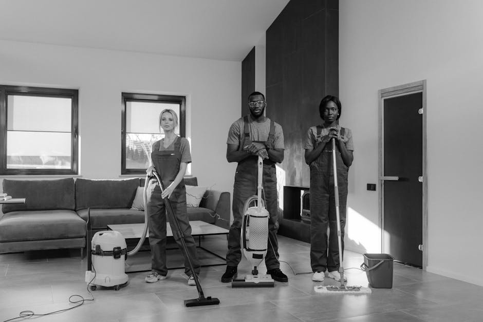 Two professional cleaners from Finchley Cleaner are performing surface cleaning and vacuuming on a light grey tiled floor in a modern living room. The room features a grey fabric sofa with cushions, p