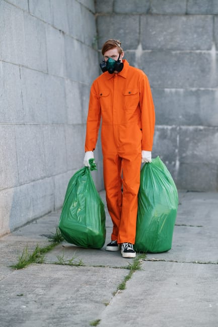 A female cleaner wearing a light blue uniform is using a commercial waste compactor outdoors, situated on a paved area near a modern building with large windows and a landscaped garden featuring trimm